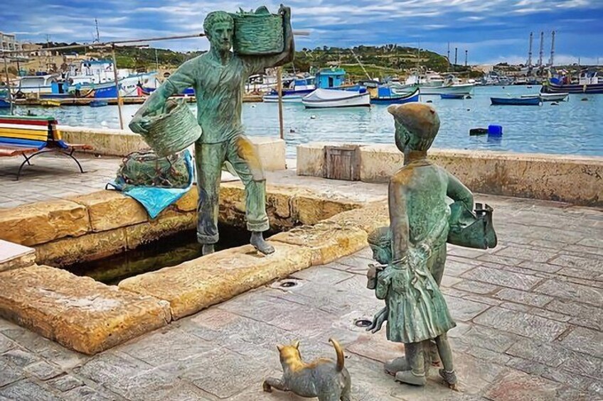 A statue of a man carrying a basket, a statue of a child, and a small dog in a harbor setting with boats in the background. 3