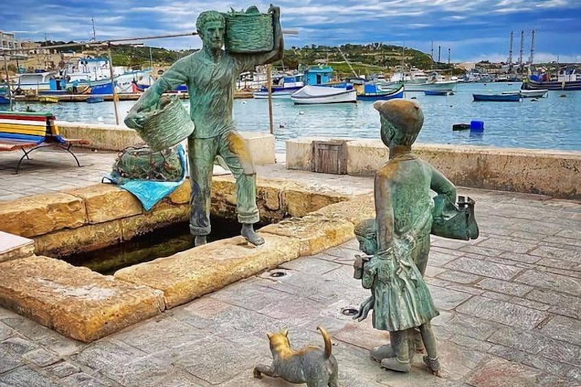 Una estatua de un hombre cargando una cesta, una estatua de un niño y un perro pequeño en un puerto con barcos al fondo. 4