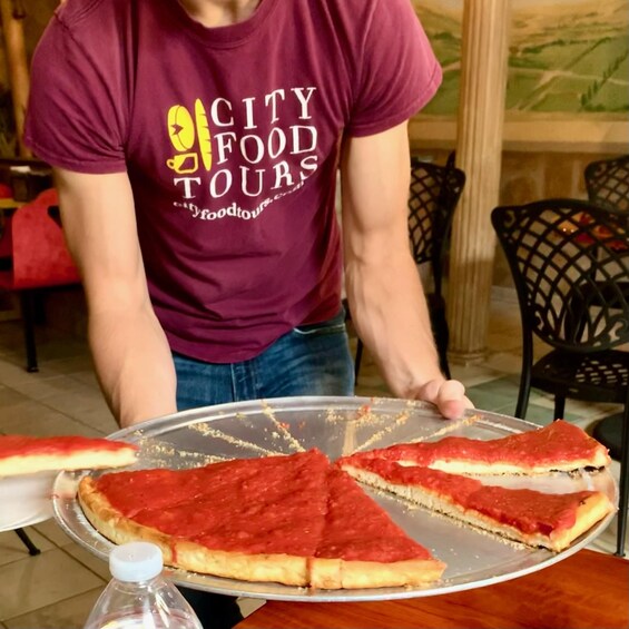 A person wearing a 'CITY FOOD TOURS' t-shirt serving a large pizza on a tray. 1
