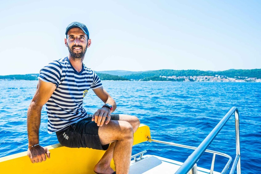 A man sitting on a yellow boat with a striped shirt and black shorts, with a coastal landscape in the background. 3
