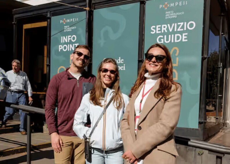 Three people standing in front of a building with 'SERVIZIO GUIDE' and 'INFO POINT' signs. 2