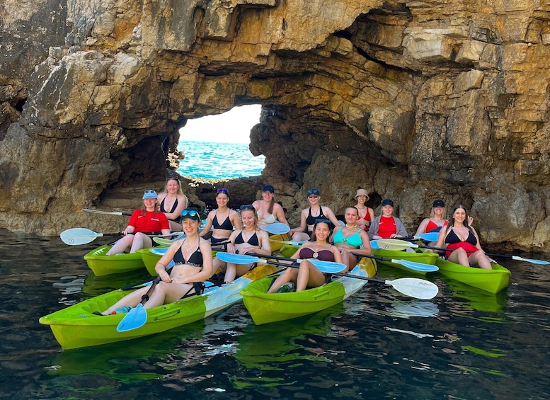 A group of people in green kayaks sitting in front of a rocky cave entrance with water. 3