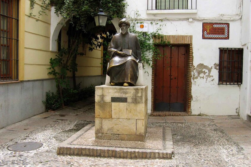 A statue of a man in traditional attire on a pedestal in a courtyard with cobblestone flooring, surrounded by buildings with a sign above a door. 3