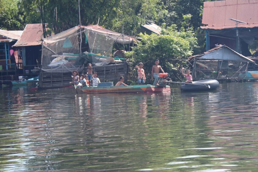 Eine Gruppe von Menschen in einem kleinen Boot auf ruhigem Wasser, umgeben von einfachen Gebäuden und viel Grün. 5