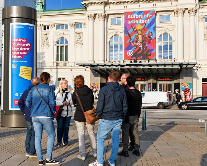 Un groupe de personnes se tient sur un trottoir, devant un bâtiment à l’architecture classique, et une grande affiche annonce des travaux de construction. 3