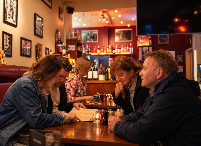 Un groupe de personnes est assis à une table dans un bar, avec un menu, des bouteilles et un téléviseur en arrière-plan. 4