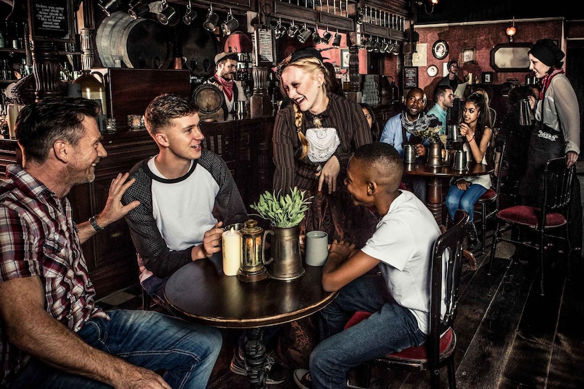 A group of people sitting around a table in a bar with various bottles and a plant on the table. 4