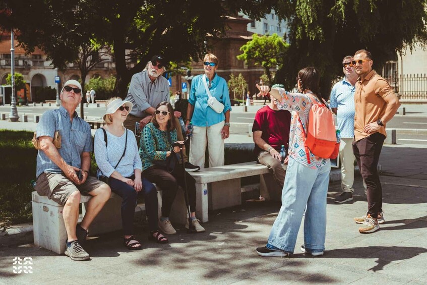 A group of people sitting and standing on a bench in a park with trees and buildings in the background. 1
