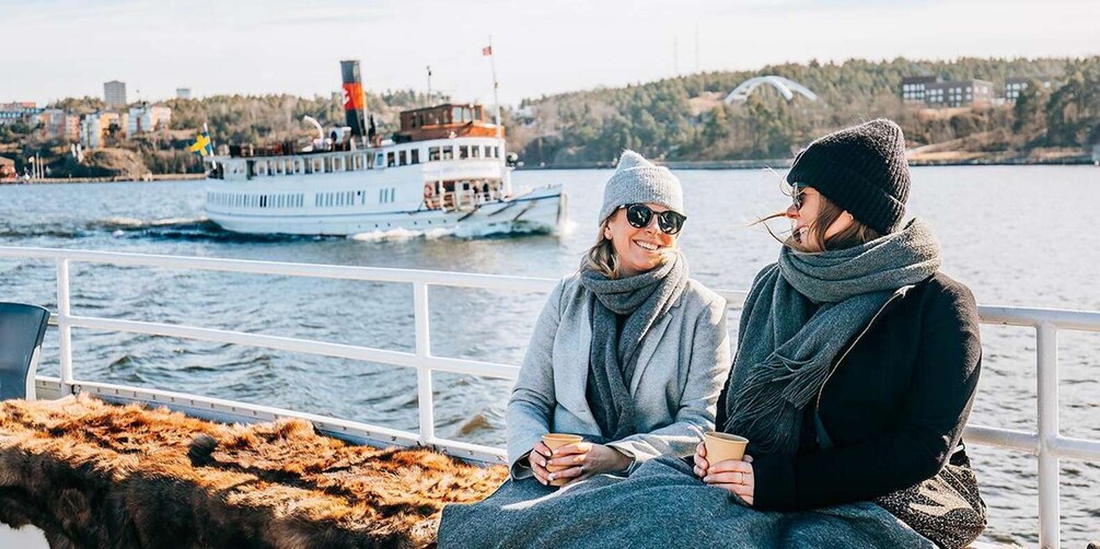 Two people sitting on a boat, holding coffee cups, with a river and a ferry in the background. 1