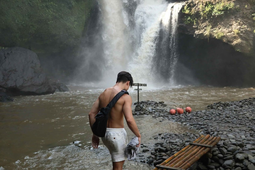 Un uomo in piedi vicino a una cascata, tiene in mano un asciugamano, con uno zaino e un segnale stradale sullo sfondo. 4