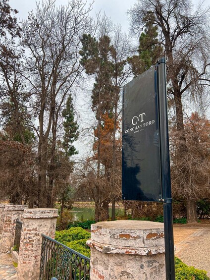 A sign with 'CT' and 'CONCHA TORO' written on it, surrounded by trees and stone structures. 3