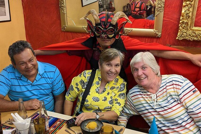 Tres personas sentadas en una mesa con una botella, un plato de comida y una máscara sobre la mesa. 1