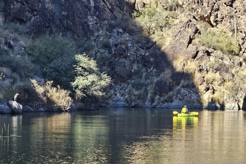 Een kajakker in een gele kajak op een kalme rivier, omgeven door rotsachtige kliffen en schaarse begroeiing. 3