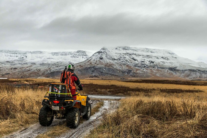 A person riding a yellow ATV on a dirt path with snow-capped mountains in the background. 1