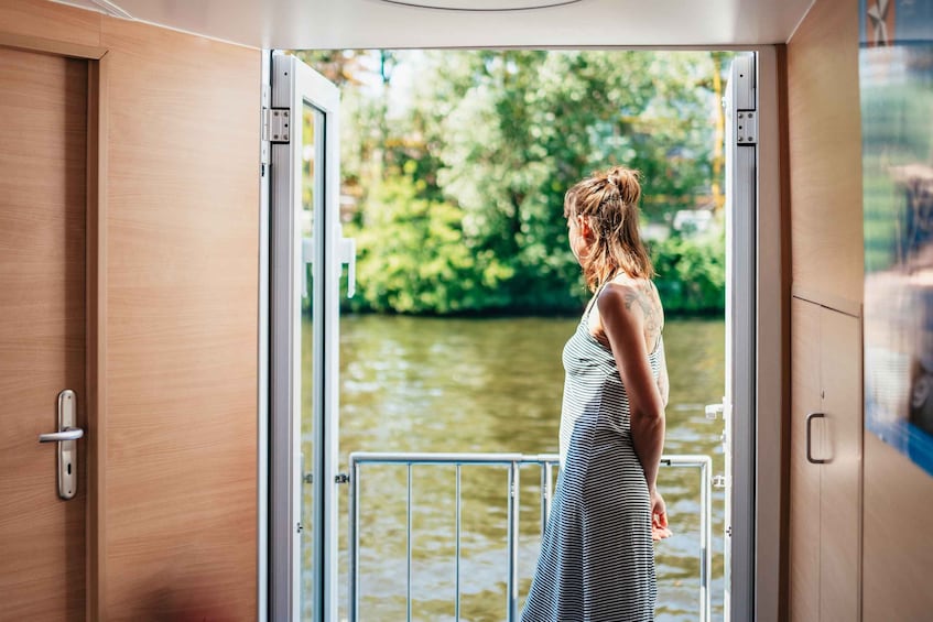 Una mujer con vestido a rayas está de pie en la puerta abierta de un bote, mirando un río con árboles al fondo. 1