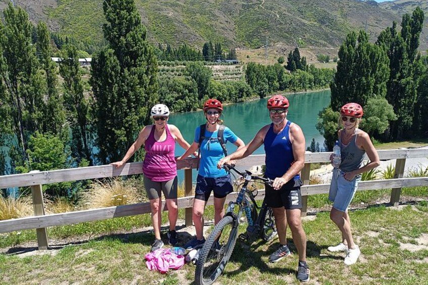 Vier personen staan bij een meer, met fietsen, en dragen helmen, met een houten omheining op de achtergrond. 2