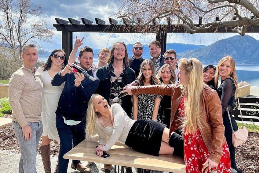 A group of people gathered around a table outdoors with a scenic lake and mountains in the background. 2