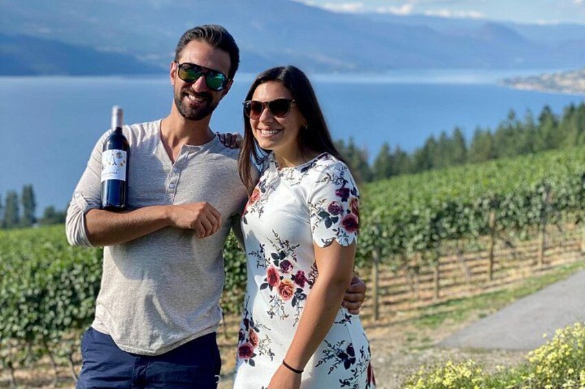 Un hombre y una mujer de pie en un viñedo, con una botella de vino, una vista panorámica de un lago y un cielo despejado. 1