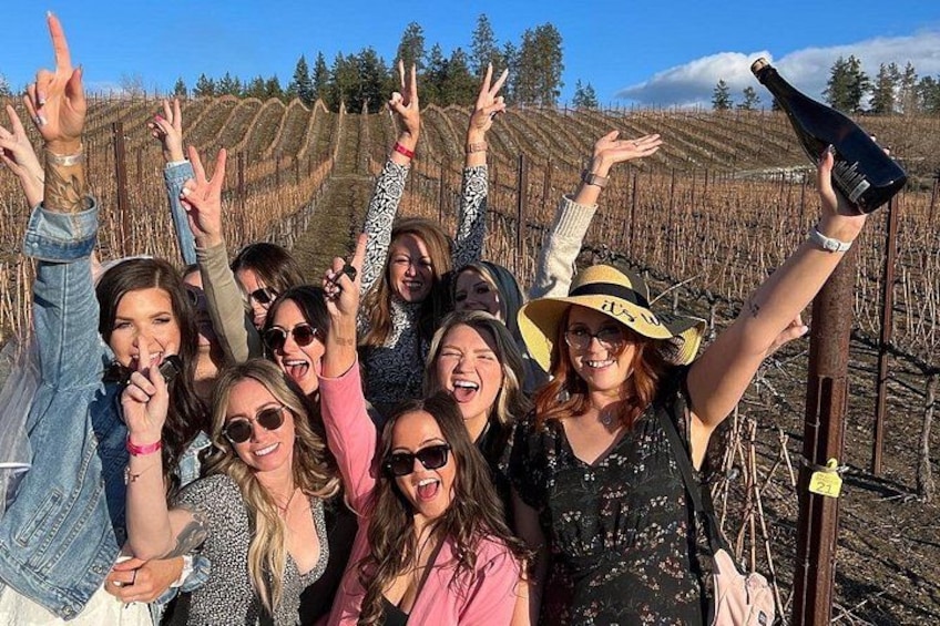 A group of people in a vineyard with raised hands, a bottle of wine, and a sign on a post. 3