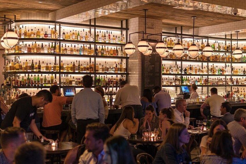 A bar with shelves of bottles, people standing and sitting, and a menu board. 4