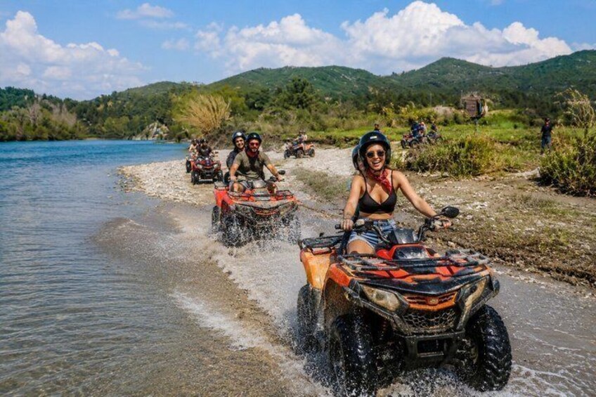 People riding ATVs along a riverbank with a mountainous landscape in the background. 1