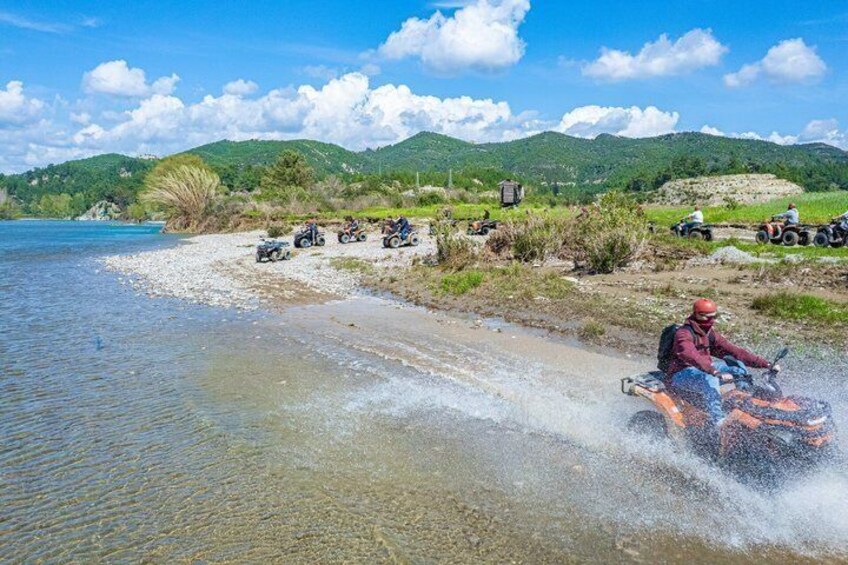 A person riding an ATV on a riverbank with other ATVs and people in the background. 4