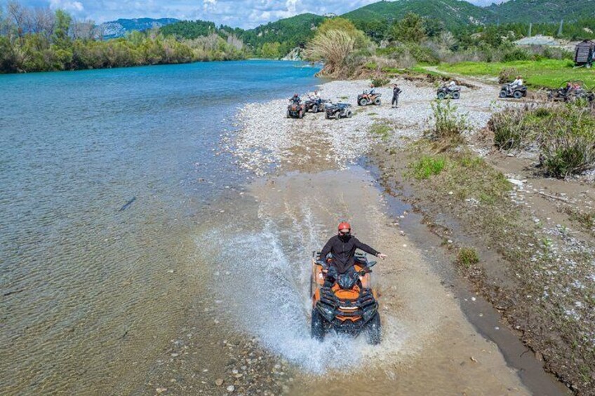 A person on a quad bike crossing a shallow river, surrounded by a group of vehicles and people on a dirt path. 3