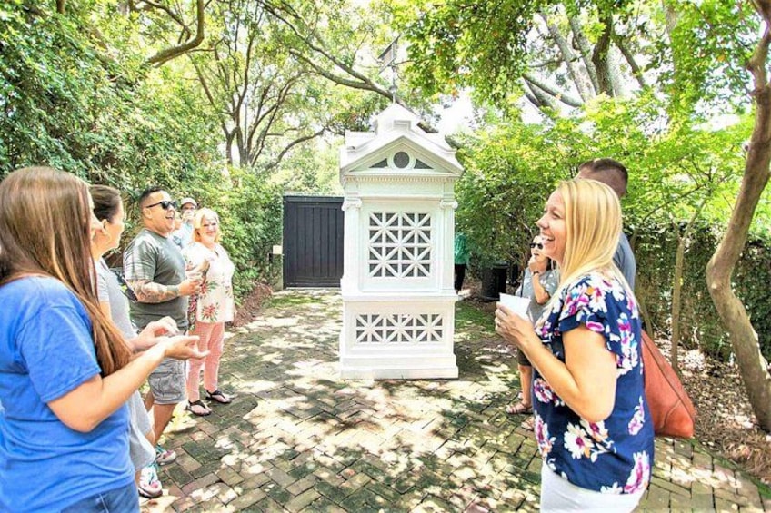 A group of people standing outdoors near a white gazebo with a lattice design, surrounded by trees and a brick pathway. 5