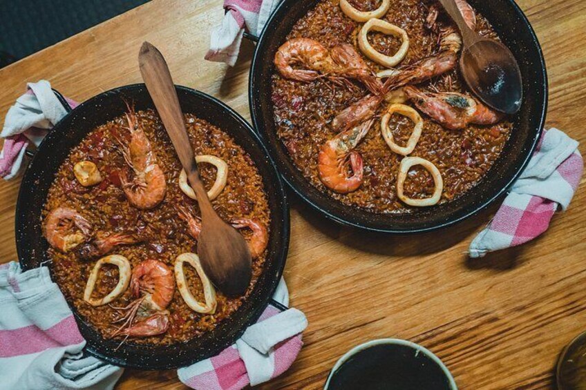 Due padelle di paella di pesce con gamberi e calamari fritti, cucchiai di legno e tovagliolo a quadretti su un tavolo di legno. 4
