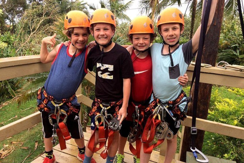 Four people wearing helmets and harnesses on a wooden bridge with greenery in the background. 4