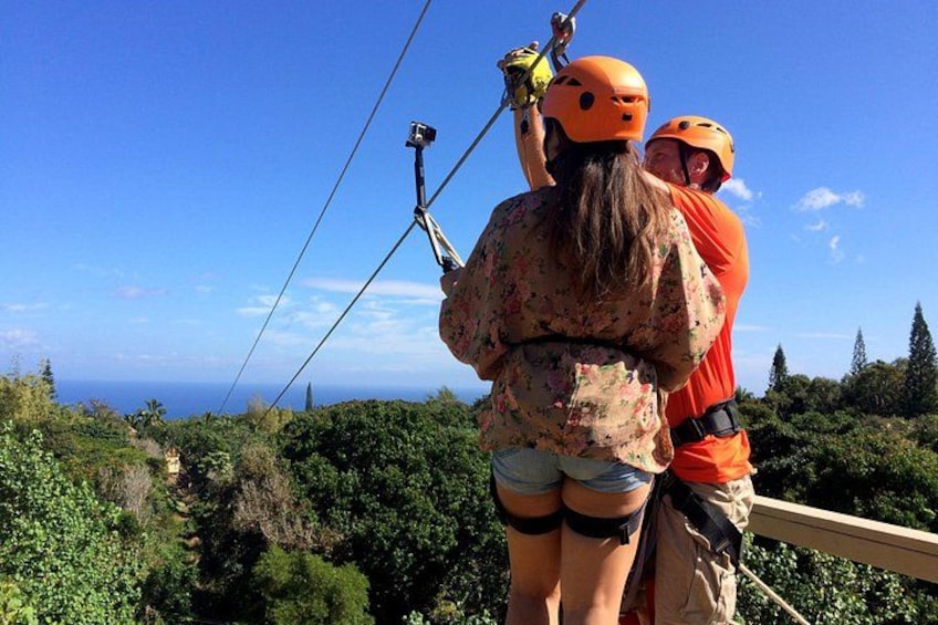 Two people on a zip line with helmets, overlooking a forested landscape and the ocean. 1