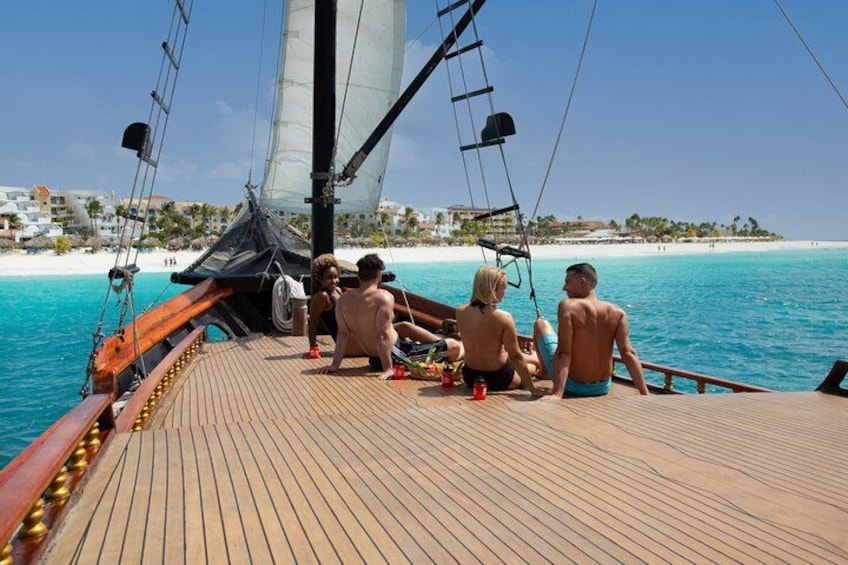 A sailboat on calm waters with a wooden deck, a clear sky, and a beach with buildings in the background. 3
