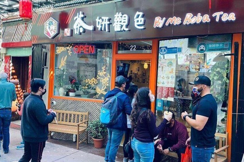 Un salon de thé avec une enseigne dans une langue étrangère, un banc en bois à l’extérieur et des gens qui se tiennent devant. 2