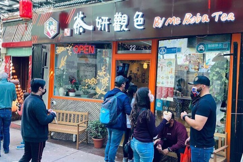 A tea shop with a sign reading "By Me Boba Tea" and a "OPEN" sign. People are gathered outside, some looking at the shop and others engaged in conversation. 2