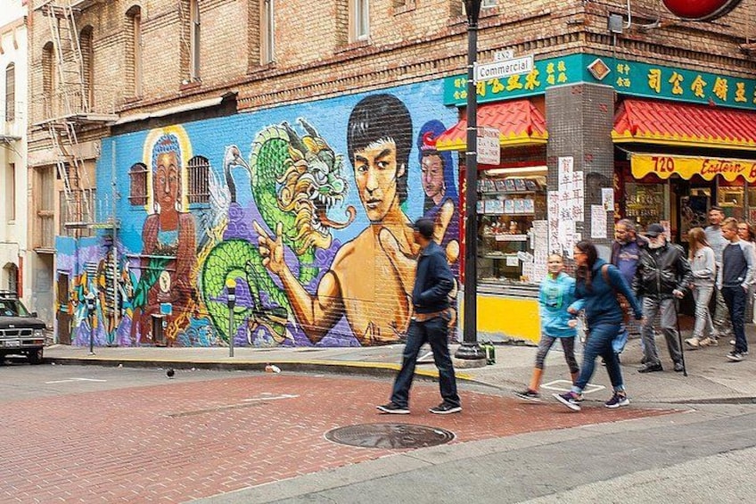 Un mural con un dragón y una persona en un edificio, una calle con peatones y una tienda con un letrero. 1