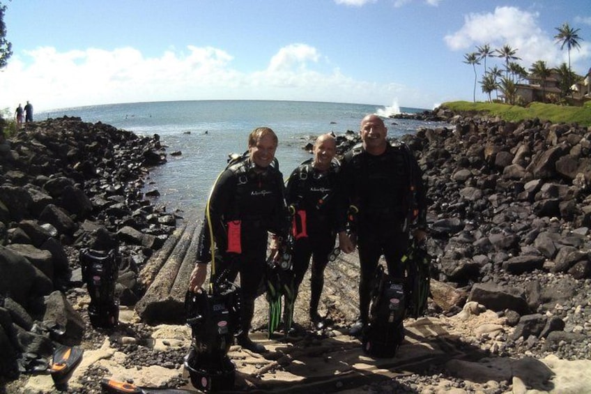 Three people in scuba diving gear standing on a rocky beach with the ocean in the background. 4