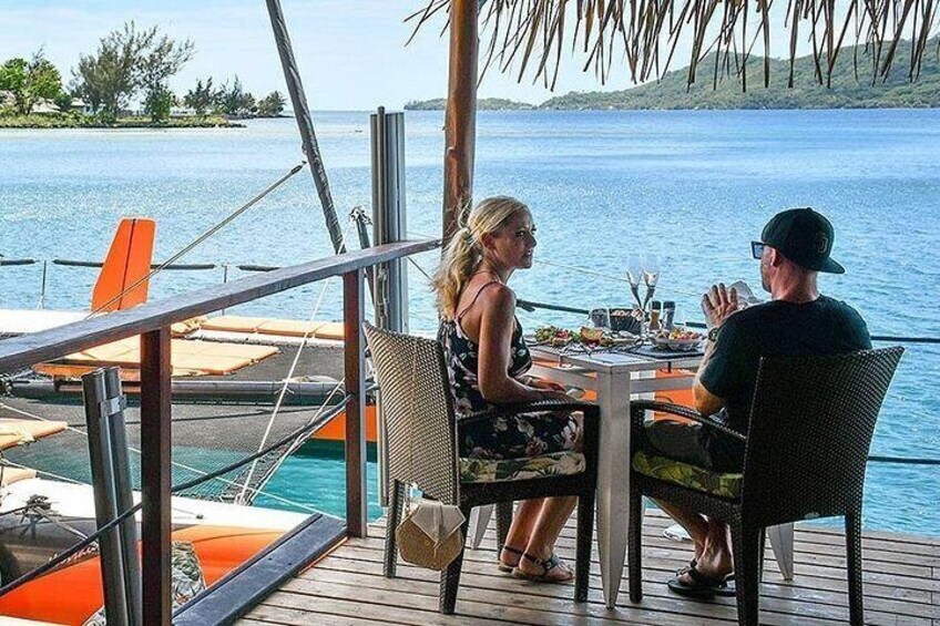 A couple dining at a table on a wooden deck with a view of the sea and boats. 2