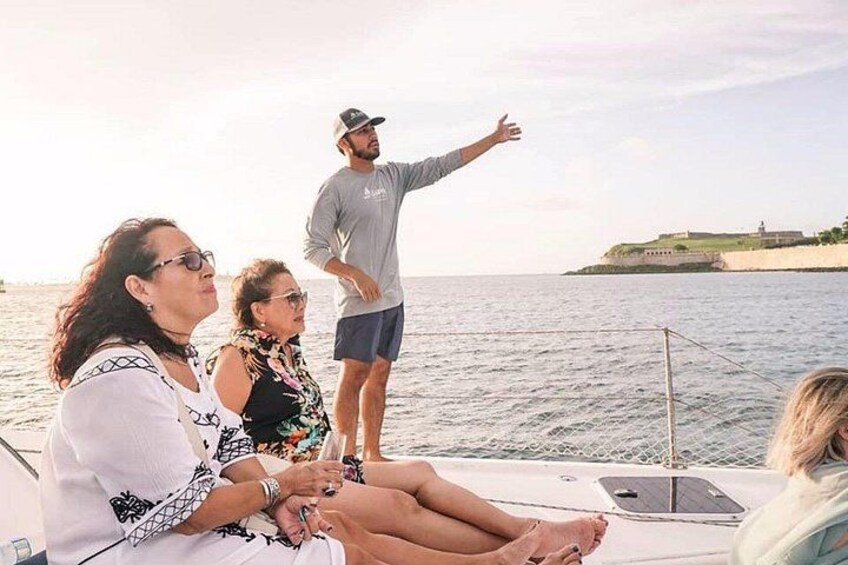 A group of people on a boat with a person standing and gesturing, a calm sea, and a distant shoreline with buildings. 5