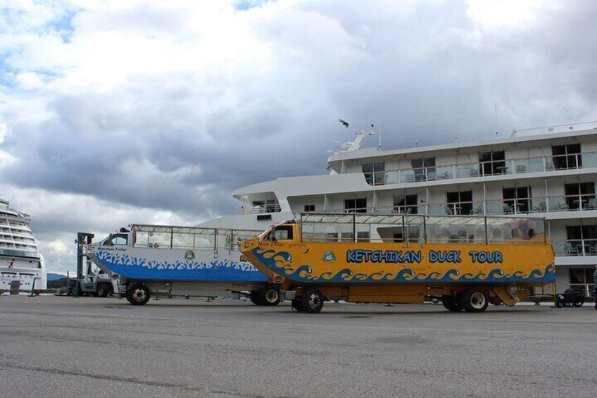 Een veerboot met een geel en blauw bord waarop 'Ketchikan Duck Tour' staat, een witte cruiseschip en een bewolkte lucht. 5