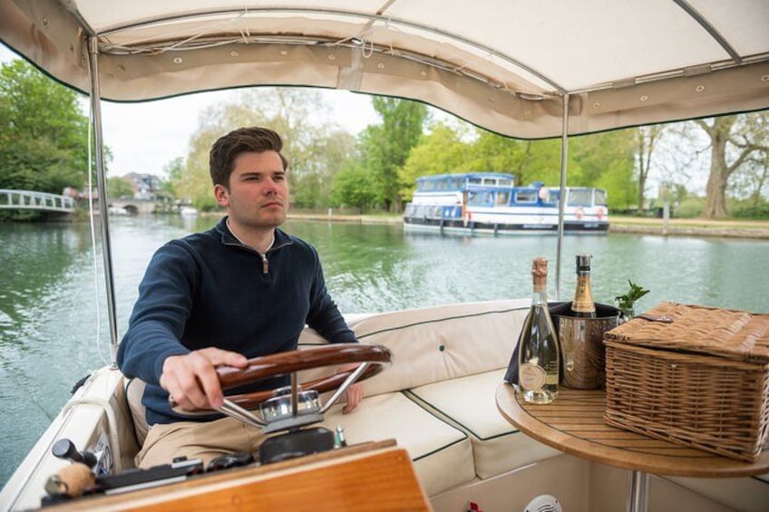 A person steering a boat on a river with a wicker basket and bottles on the deck. 1