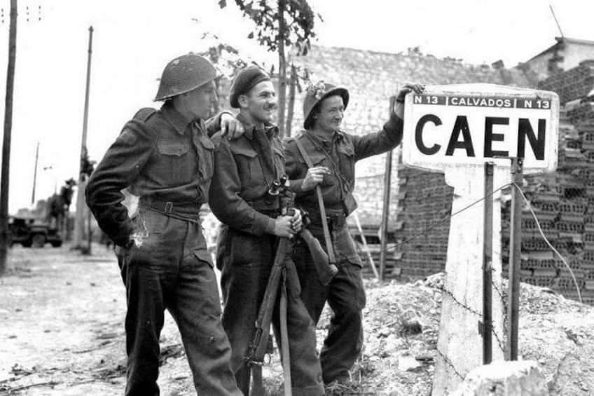Soldiers standing next to a sign that reads 'CAEN' with a road sign in the background. 4