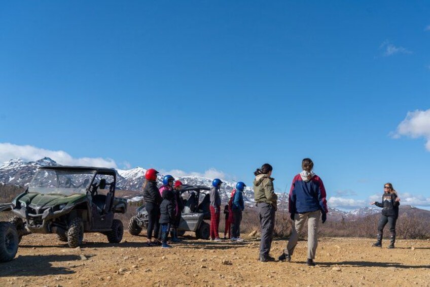 Un grupo de personas está de pie en un camino de tierra, con un vehículo y un paisaje montañoso al fondo. 3