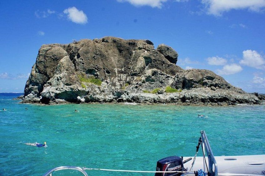 A rocky island surrounded by turquoise water, a boat with a visible engine, and a clear blue sky. 2