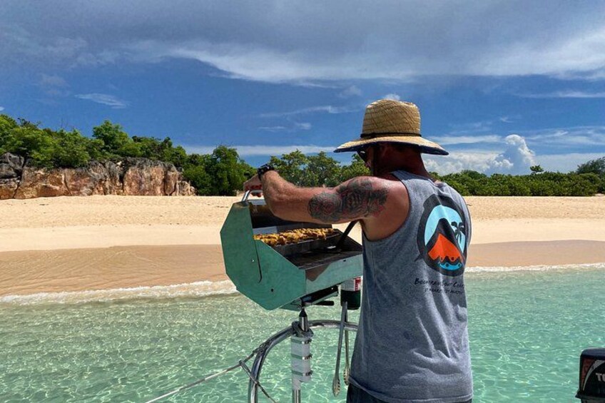 A person grilling on a boat near a beach with clear water and a rocky shoreline. 2