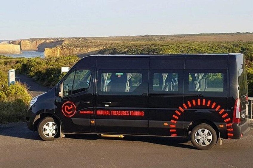 A black van with 'Natural Treasures Touring' written on the side parked on a road with a coastal landscape in the background. 2