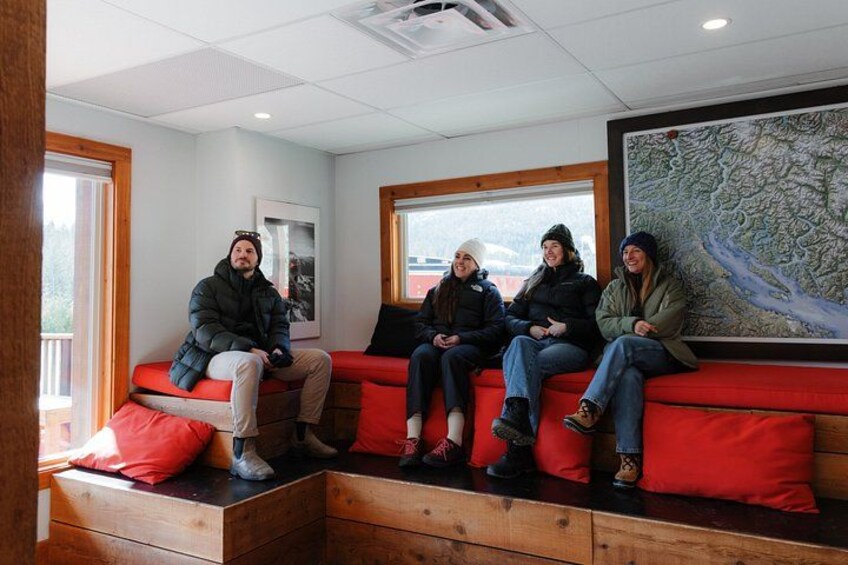 Un groupe de personnes est assis sur un banc rouge, dans un espace intérieur agrémenté d’éléments en bois et orné d’une grande carte au mur. 3