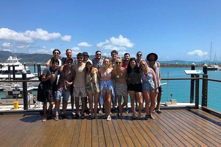 Un gruppo di persone su un molo di legno, vicino a un porto turistico con barche e cielo sereno. 2