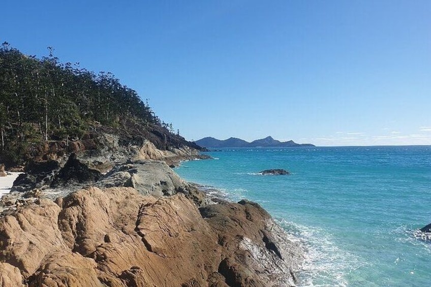Paesaggio costiero con scogliere rocciose, acqua turchese limpida e montagne in lontananza. 4