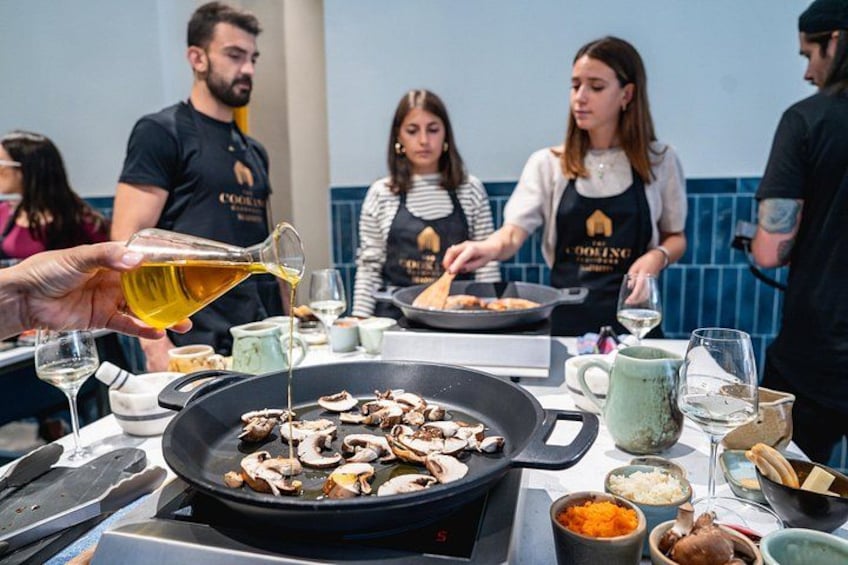 Una clase de cocina con participantes preparando alimentos, una sartén con champiñones y utensilios de cocina sobre una mesa. 5