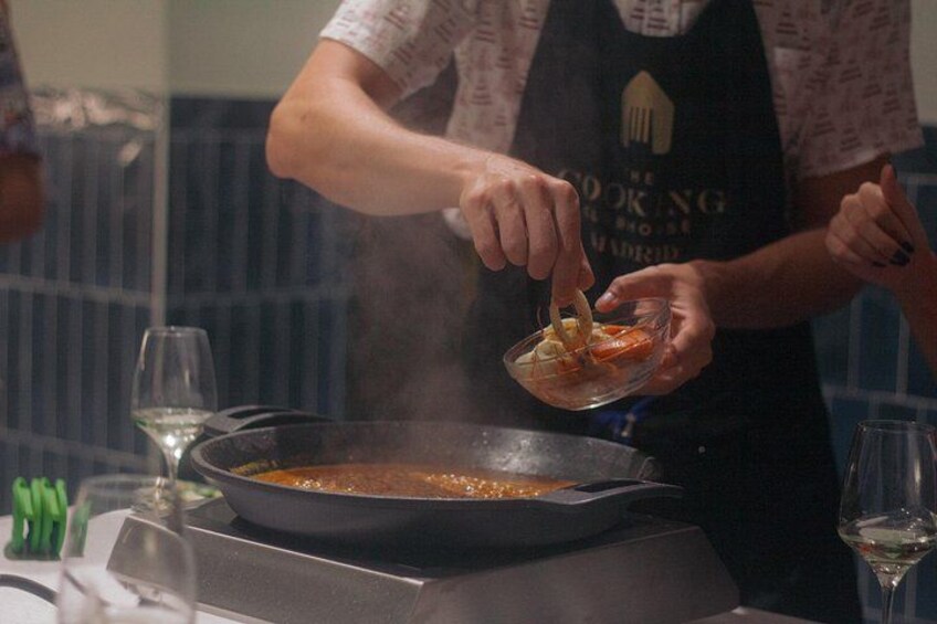 Una persona cocina con una sartén sobre la estufa, una copa de vino y una botella de vino en la mesa. 2